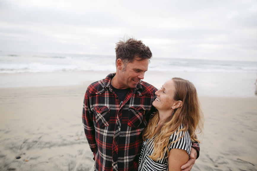 A smiling man and woman embracing on a beach with waves in the background, representing the 'Adults' category.