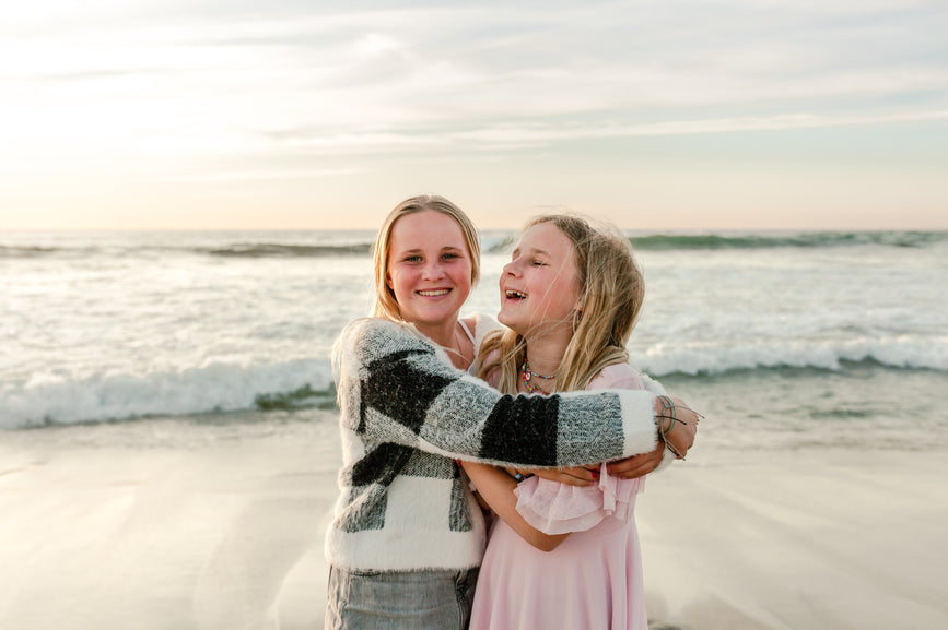 Two smiling young girls, embracing on a beach with waves in the background, representing the 'Teens and Tweens' category.