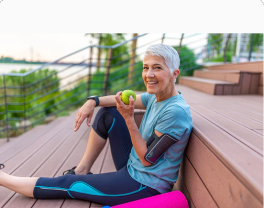 A smiling older woman with short grey hair, wearing athletic clothing, sitting on wooden steps outdoors and eating a green apple. A pink yoga mat is beside her.